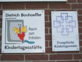 Es ist das Logo der evangelischen Dietrich-Bonhoeffer-Kindertagesstätte zu sehen. Dieses setzt sich aus grauen Rechtecken und einem gelb/roten Schmetterling mit entsprechenden Schriftzug zusammen.