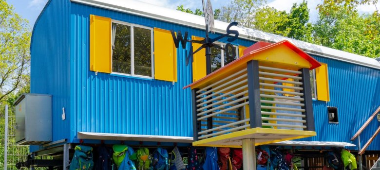 Im Vordergrund ein Vogelhaus aus Kunststoff. Dahinter ein blauer Bauwagen mit gelben Fensterläden an dem Kinderjacken hängen.