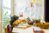 Raum einer Kita zwei Stoffpuppen sitzen an einem Tisch und es sieht aus als würden sie spielen und basteln auf dem Tisch steht ein Ständer mit Scheren ein Farbkreis liegt dort und ein buntes Blatt Papier