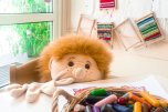 Raum einer Kita. Links ein Fenster zum Garten, davor sitzt eine große Handpuppe am Tisch. Es sieht aus als würde die Puppe malen. Auf dem Tisch liegt ein rot bemaltes Blatt und ein Korb mit bunten Wachsmalstiften.