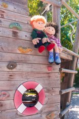 Zwei Stoffpuppen sitzen auf dem Holzschiff im Kletterbereich des Kita Gartens der Maria-Jockel-Kita. Im Hintegrund sieht man das Grün eines Baums.