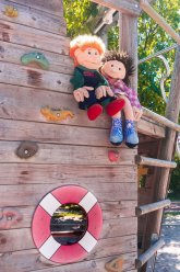Zwei Stoffpuppen sitzen auf dem Holzschiff im Kletterbereich des Kita Gartens der Maria-Jockel-Kita. Im Hintegrund sieht man das Grün eines Baums.