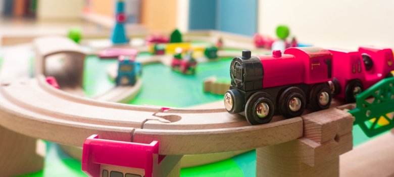 Nahaufnahme einer Holzeisenbahnbrücke auf der eine rote Spielzeuglock von rechts ins Bild fährt im Hintegrund unscharf die bunte Spiellandschaft