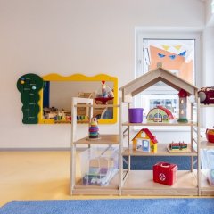 Man sieht eine Sandfläche des Außenbereichs, Darauf steht ein Klettergerüst in Form eines kleinen Busses und eine Rutsche.