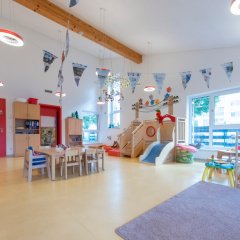 Es ist ein bunter Gruppenraum zu sehen. In ihm steht eine Tischgruppe mit Stühlen, eine Hochebene und rechts im Bild ist ein Regal mit Spielzeugen zu sehen.