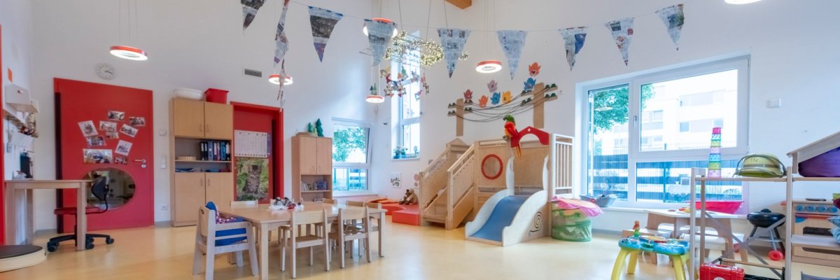 Es ist ein bunter Gruppenraum zu sehen. In ihm steht eine Tischgruppe mit Stühlen, eine Hochebene und rechts im Bild ist ein Regal mit Spielzeugen zu sehen.