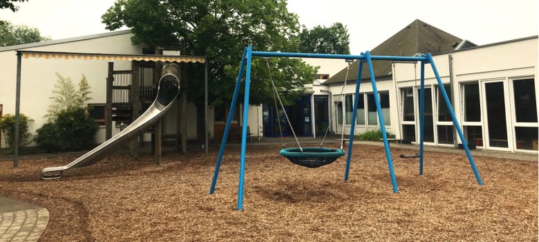 Ein Teil des Außengeländes der Kita ist sichtbar. Man sieht eine Rutsche und eine Korbschaukel