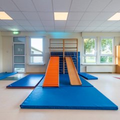 In der Raummitte sind mehrere Turnmatten auf dem Boden. An der Sprossenwand ist eine Bank eingeharkt, sodass man darauf runterrutschen kann.
