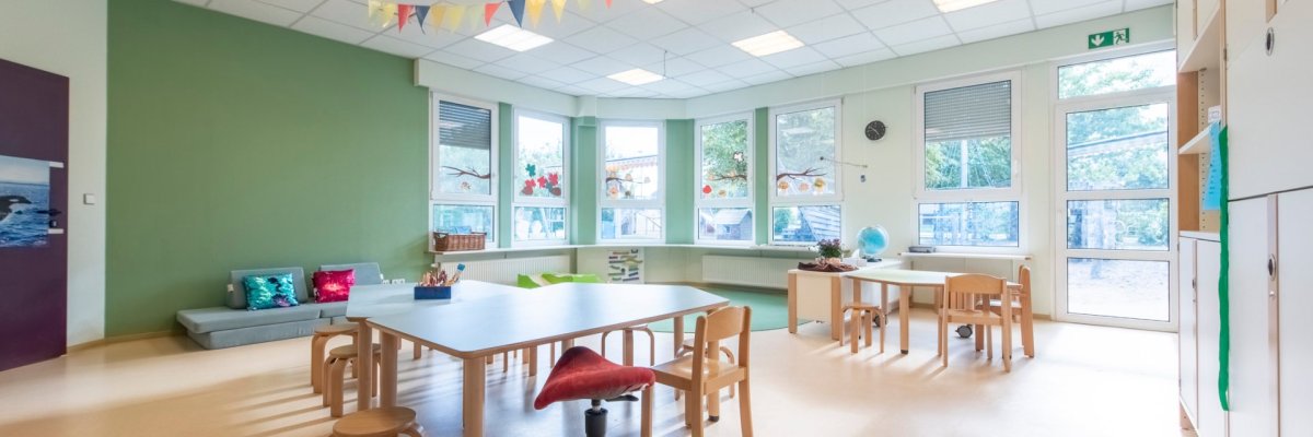 Es ist einer der Gruppenräume der Einrichtung zu sehen. Im Raum stehen Tischgruppen. Durch die helle Fensterfront, wirkt der Raum einladend.