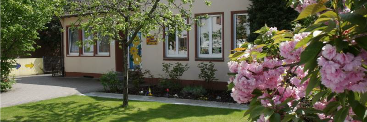 Es ist die Außenfassade der evangelischen MArtin-Luther Kita zu sehen mit einer grünen Wiese und einem rosa blühenden Busch vor dem Gebäude.