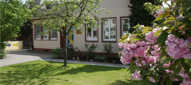 Es ist die Außenfassade der evangelischen MArtin-Luther Kita zu sehen mit einer grünen Wiese und einem rosa blühenden Busch vor dem Gebäude.