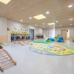 Spielflur der Bewegungskita Rheinakrobaten ist zu sehen. Links im Bild sieht man einen Kletterbogen und große Plüschteddybären. An der linken Seite sind die Umkleiden für die Kinder. Mittig im Bild ist ein HIndernisparcour aufgebaut - bestehend aus Balancierübung, Schleichtunnel und Kletterdreieck mit Rutsche.