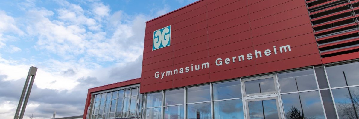 Schräg von rechts nach links verlaufender Blick auf die Front des Gymnasiums mit Hauptblick auf die Verglasung der Mensa und der großen Beschriftung des Gebäudes.