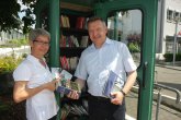 Links im Bild Frau Hildegard Bolenz rechts Herr Bürgermeister Peter Burger bei der Einweihung der öffentlichen Bücherzelle in Gernsheim, welche man in türkiser Farbe hinter den beiden sehen kann. Beide Personen haben Bücher in der Hand.