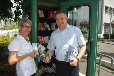 Links im Bild Frau Hildegard Bolenz rechts Herr Bürgermeister Peter Burger bei der Einweihung der öffentlichen Bücherzelle in Gernsheim, welche man in türkiser Farbe hinter den beiden sehen kann. Beide Personen haben Bücher in der Hand.