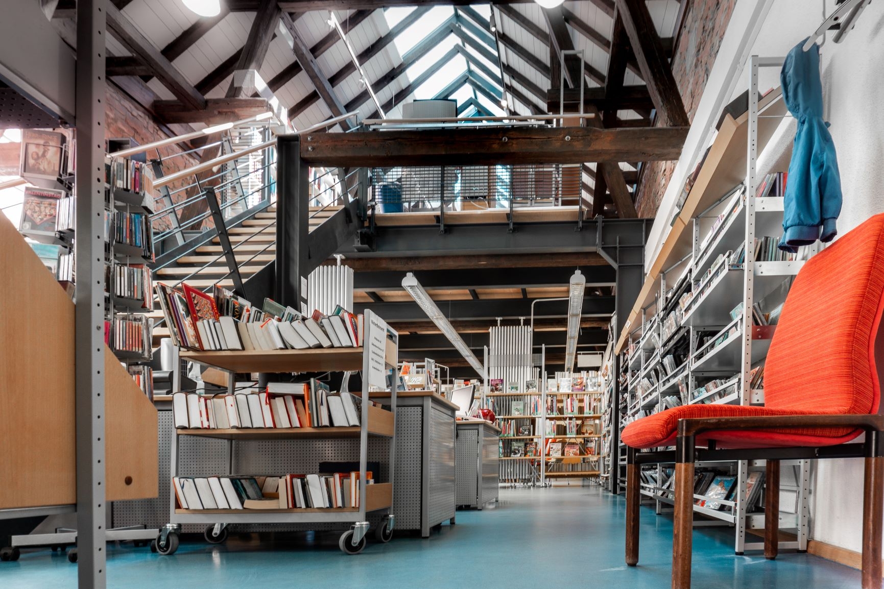 Froschperspektive vom Eingang der Bücherei aus ins Erdgeschoss. Ein oranger Polsterstuhl steht rechts unter der Garderobe an der eine blaue Kinderjacke hängt. Man sieht die Medienregale rechts. Mittig die Arbeitsplätze des Teams und links die Treppe ins OG. Im oberen Bildrand sieht man das Fachwerk des Obergeschosses.