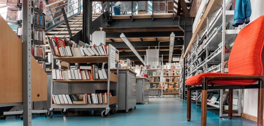Froschperspektive vom Eingang der Bücherei aus ins Erdgeschoss. Ein oranger Polsterstuhl steht rechts unter der Garderobe an der eine blaue Kinderjacke hängt. Man sieht die Medienregale rechts. Mittig die Arbeitsplätze des Teams und links die Treppe ins OG. Im oberen Bildrand sieht man das Fachwerk des Obergeschosses.