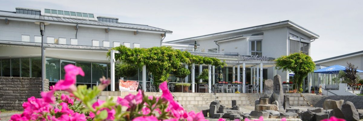 Die Stadthalle Gernsheim von vorne im Vordergrund unscharf eine Pflanze mit pinken Blüten über die hinweg fotografiert wurde.