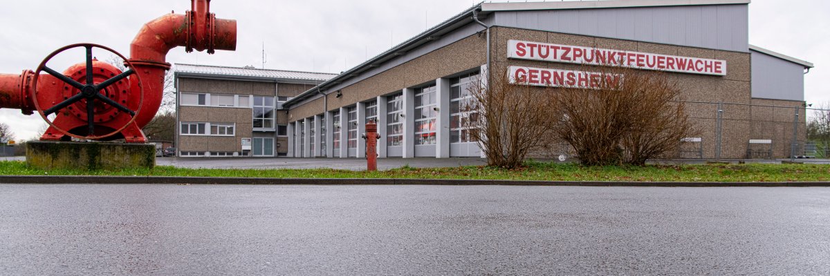 Stützpunkfeuerwache Gernsheim. Man sieht die Fahrzeughalle rechts und das Bürogebäude mittig. Am linken Bildrand ist im Vordergrund ein großes rotes Element einer alten Pumpe, welches dort dekorativ aufgestellt wurde.