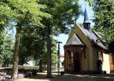 Man sieht die Kapelle Maria Einsiedel rechts im Bild in hellem Gelb angelegt mit Sandsteinportal und einem kleinen schiefergedeckten Glückentürmchen in der Mitte des Dachs links davor sieht man Bäume die die Sitzbänke um den Außenaltar einrahmen es scheint die Sonne
