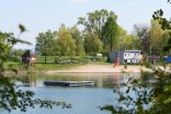 Badesee umringt von Bäumen. In der Mitte ist ein Schwimmsteg. Am Sandstrand befindet sich ein Spielturm mit Rutsche. Dahinter Graublaues Kioskgebäude mit Sonnenschirmen. Am linken Bildrand befindet sich eine rote Holzhütte mit Spitzdach.