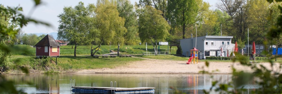 Badesee umringt von Bäumen. In der Mitte ist ein Schwimmsteg. Am Sandstrand befindet sich ein Spielturm mit Rutsche. Dahinter Graublaues Kioskgebäude mit Sonnenschirmen. Am linken Bildrand befindet sich eine rote Holzhütte mit Spitzdach.