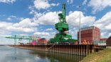 Wasserfläche des Hafens. Oberhalb der Spundwand ein grüner Kran mit vielen Containern. Im Hintergrund ein grüner Brückenkran.