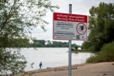 Im Vordergrund ein Warnschild vor dem Baden im Rhein. Im Hintergrund der Rhein mit Sandstrand. Am Bildrand sind Bäume und Sträucher zu sehen.