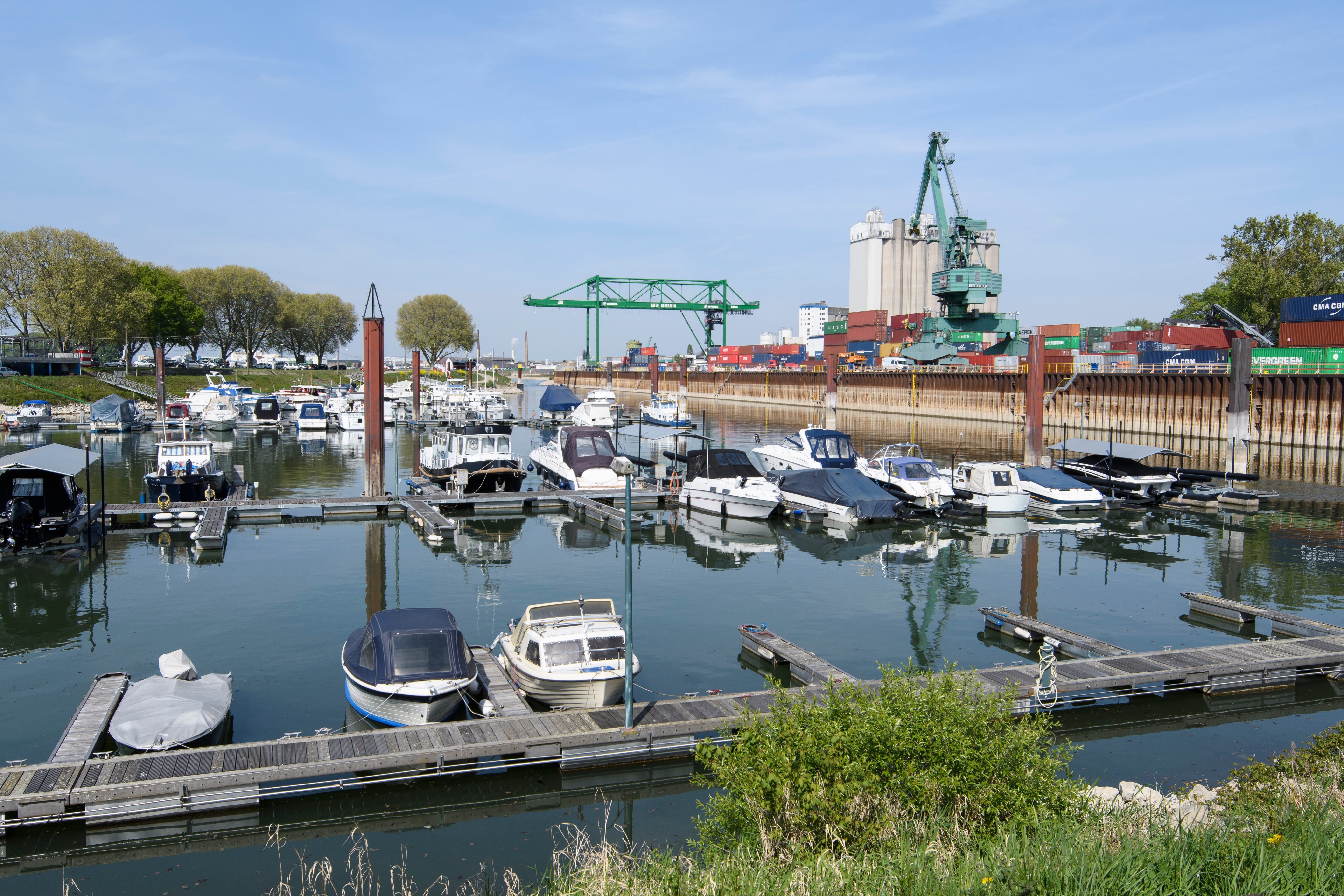 Im Vordergrund liegen an Stegen festgemachte Sportboote im Hafen. Rechts im Hintergrund ist ein Grüner Brückenkran über diversen Containern zu sehen. Links befindet sich die mit Bäumen bestandene Hafenspitze.