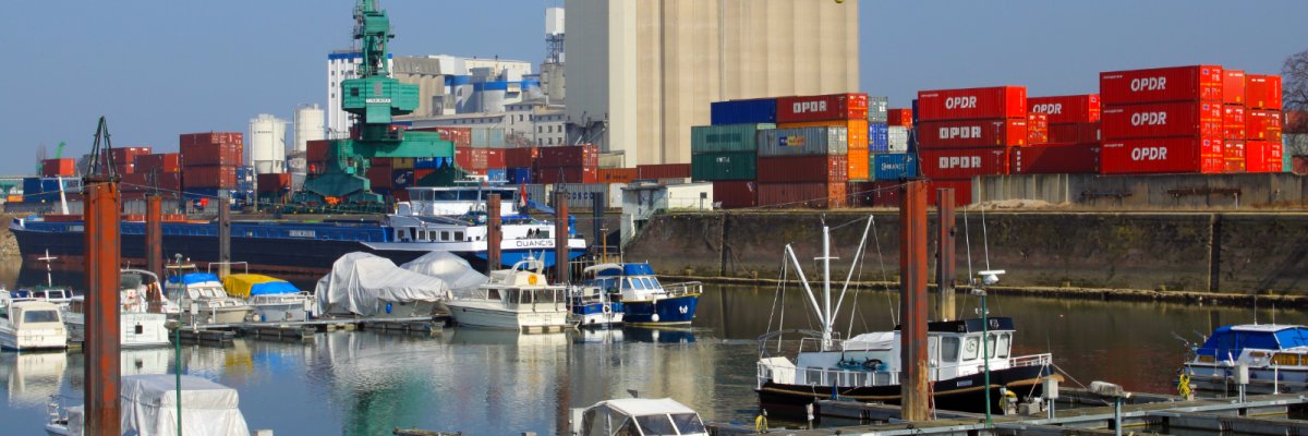 Im Vordergrund liegen an Stegen festgemachte Sportboote im Hafen. Dahinter liegt ein Tankmotorschiff mit schwarzem Rumpf. Im Hintergrund ist ein Grüner Brückenkran über diversen Containern zu sehen. 