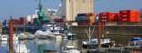 Im Vordergrund liegen an Stegen festgemachte Sportboote im Hafen. Dahinter liegt ein Tankmotorschiff mit schwarzem Rumpf. Im Hintergrund ist ein Grüner Brückenkran über diversen Containern zu sehen.