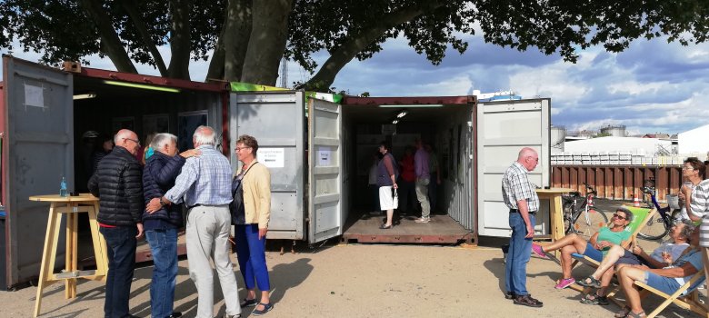 Auf der Kiesfläche der Hafenspitze stehen Liegestühle und Stehtische aus Holz an denen sich mehrere Menschen befinden. Im Hintergrund stehen zwei dunkelrote offene Frachtcontainer unter zwei großen Bäumen. In beiden Containern schauen sich Menschen Pläne an.