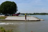 In der Mitte befindet sich die Hafenspitze von Gernsheim umgeben von Wasser des Rheins. In der Mitte der Hafenspitze befindet sich auf einem Betonsockel ein altes Fischerboot mit Namen Hannelore. Links davon stehen zwei große Bäume unter denen drei Autos parken.