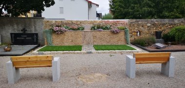 Farbig bepflanzte Sternenkindergrabstätte auf dem Gernsheimer Friedhof. Dahinter ist die alte Friedhofsmauer. Im Vordergrund mit Blick auf die Grabstätte sind zwei Bänke mit Betonsockel und Sitzfläche aus Holz.