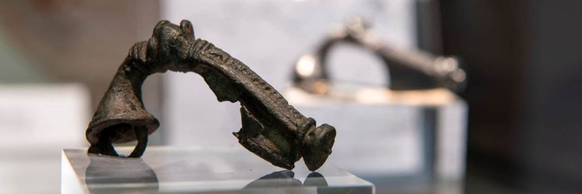 Eine antike römische Gewandfibel aus dem Bestand des Museums der Schöfferstadt Gernsheim ist scharf links im Vordergrund zu sehen. Durch die lange Zeit im Boden ist sie fast schwarz geworden. Im Hintergrund ist eine weitere Fibel unscharf abgebildet.