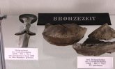 Tischvitrine mit Funden aus der Bronzezeit. Links liegt ein silbergrauer Mörigerschwertknauf von circa 1200 bis 800 vor Christus. Die Klinge ist ungefähr eine Hand breit unter dem Knauf abgebrochen und nicht mehr vorhanden. Der Knauf ist am Ende breiter als am Griff. Rechts im Bild liegen braune Scherben aus der gleichen Zeit.