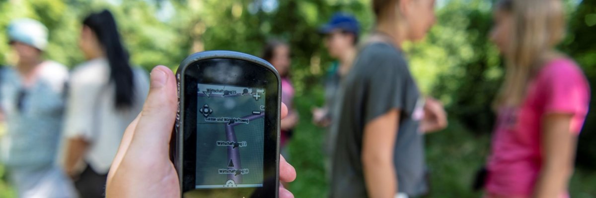 Eine Kinderhand die ein GPS Gerät fürs Geocachen hält. Im Hintegrund unscharf Kinder in weißer grauer und pinker Kleidung.