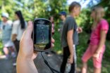 Eine Kinderhand die ein GPS Gerät fürs Geocachen hält. Im Hintegrund unscharf Kinder in weißer grauer und pinker Kleidung.
