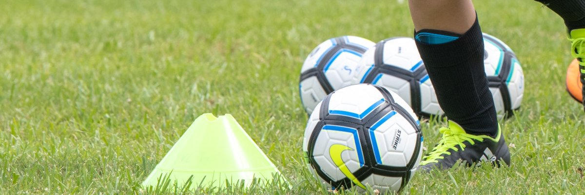 rechts im Bild Kinderbeine in Fußballkleidung bei  dribbeln eines Fußballs auf Rasen um gelbe Markierungen herum