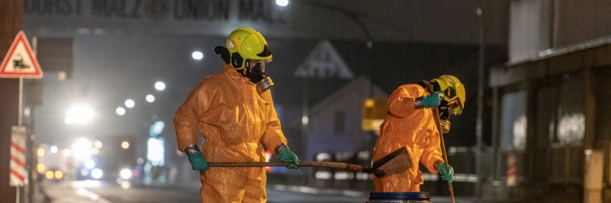 Zwei Feuerwehrleute in gelben Schutzanzügen reinigen die Straße auf Höhe der Solvadis nach einem Gefahrstoffunfall mit Besen und Schaufel und füllen die gebundenen Substanzen in ein blaues Faß.