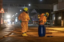 Zwei Feuerwehrleute in gelben Schutzanzügen reinigen die Straße auf Höhe der Solvadis nach einem Gefahrstoffunfall mit Besen und Schaufel und füllen die gebundenen Substanzen in ein blaues Faß.