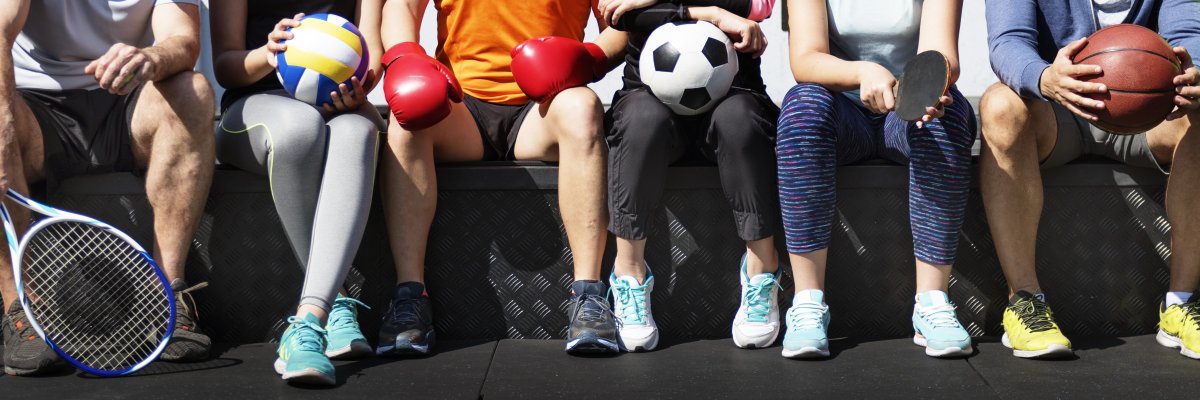 Oberkörper und Beine von sechs sitzenden Menschen. Jede hält Sportutensilien. Von links nach rechts: Tennisschläger, Volleyball, Boxhandschuhe, Fußball, Tischtennisschläger, Basketball
