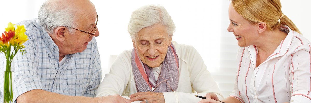 Eine jüngere Frau bespricht mit einer Seniorin und einem Senioren Unterlagen. Alle lächeln.