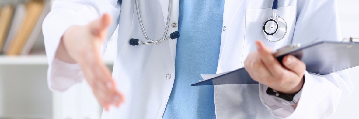 Oberkörper einer Person mit hellblauem Oberteil unter einem geöffneten weißen Arztkittel. Ein Stethoskop hängt um den Hals. Die linke Hand hält ein Klemmbrett. Die Rechte ist zum Hände schütteln ausgestreckt.
