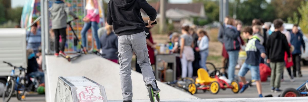 Kind mit Helm und Roller mit dem Rücken zur Kamera steht auf einem Element der Skaterbahn am Rhein. Im Hintergrund sieht man unscharf den Auszeitbus mit vielen Kindern und Jugendlichen davor.