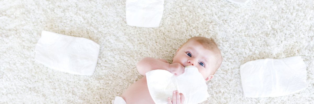 Nur mit Windel bekleidetes Baby liegt auf einem weißen Teppich. Es hält eine Windel in der Hand. Um das Baby herum liegen 5 Windeln.