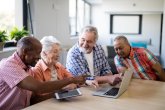 Drei Senioren und eine Seniorin sitzen an einem Tisch auf dem ein Tablet und ein Laptop steht und unterhalten sich.