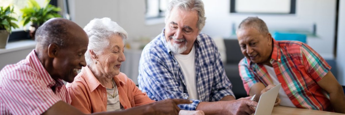 Drei Senioren und eine Seniorin sitzen an einem Tisch auf dem ein Tablet und ein Laptop steht und unterhalten sich.