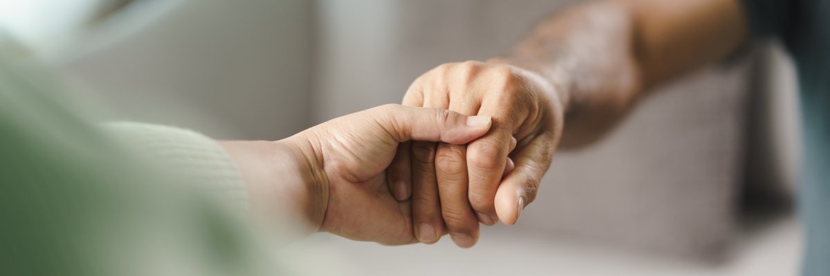Auf dem Bild sind zwei Personen zu sehen, die sich an den Händen halten. Eine Hand umfasst die andere sanft – eine Geste von Nähe, Unterstützung oder Trost. Der Hintergrund ist unscharf.