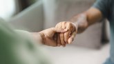 Auf dem Bild sind zwei Personen zu sehen, die sich an den Händen halten. Eine Hand umfasst die andere sanft – eine Geste von Nähe, Unterstützung oder Trost. Der Hintergrund ist unscharf.